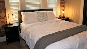 Una cama cuidadosamente hecha con edredón blanco y almohada estampada, flanqueada por dos mesitas de noche con lámparas, en una habitación con paredes de ladrillo y una ventana con persianas.