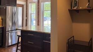 A kitchen with dark cabinets, stainless steel appliances, a hanging pendant light, and a brick wall. - 131 Cedar 2 · Chic 2BR Urban Retreat | Downtown Kalamazoo (Kalamazoo)