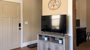 A living room with a flat-screen TV on a wooden stand, a door, and a wall-mounted artwork. - 131 Cedar 2 · Chic 2BR Urban Retreat | Downtown Kalamazoo (Kalamazoo)