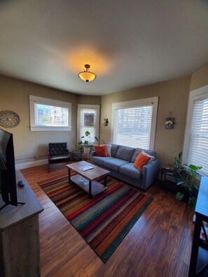 A living room with a sofa, coffee table, and a television. There are windows with blinds, a ceiling light, and a plant. - 131 Cedar 2 · Chic 2BR Urban Retreat | Downtown Kalamazoo (Kalamazoo)