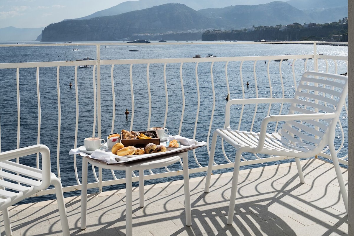 A balcony with white chairs and a table with pastries, overlooking a body of water and distant mountains.