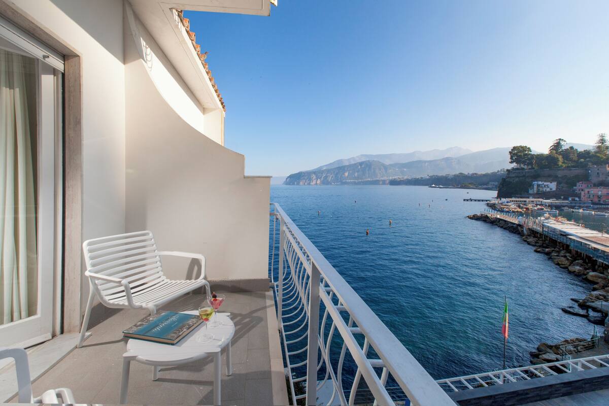 A balcony with a white chair, a small table, and a view of a calm sea with boats and distant mountains.