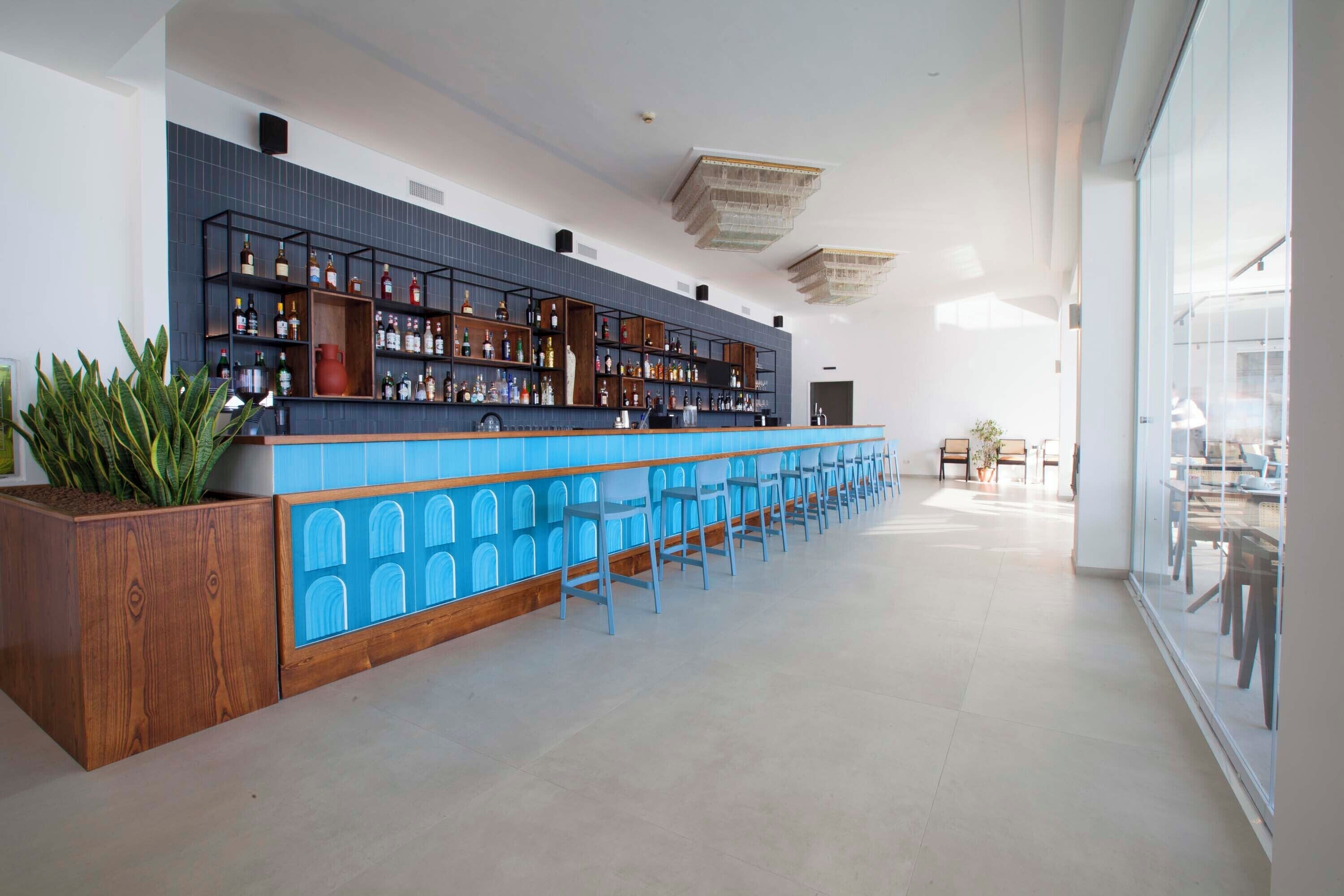 A modern bar area with a long counter, blue chairs, and a variety of bottles.