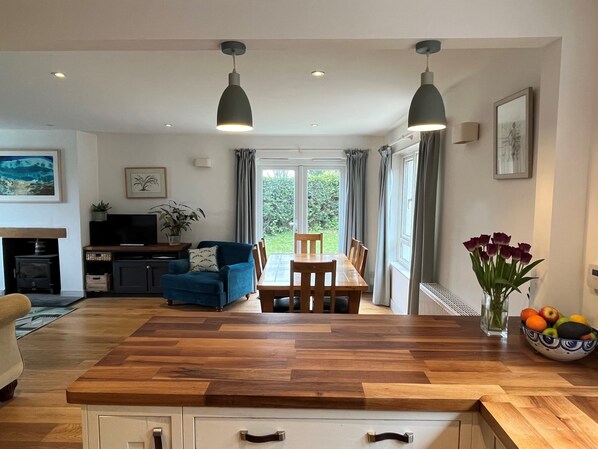A modern kitchen with a wooden countertop, two pendant lights, a dining table with chairs, a blue sofa, a TV, and a fireplace.