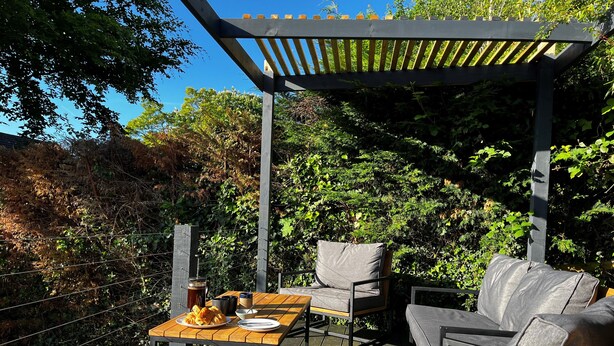 Eine Terrasse im Freien mit einem Holztisch, zwei grauen Sitzgelegenheiten mit Polstern und einer Pergola.