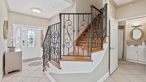 Un hall d’entrée spacieux avec un grand escalier, une porte blanche à panneaux de verre et une salle de bain moderne dotée d’un grand miroir rond.