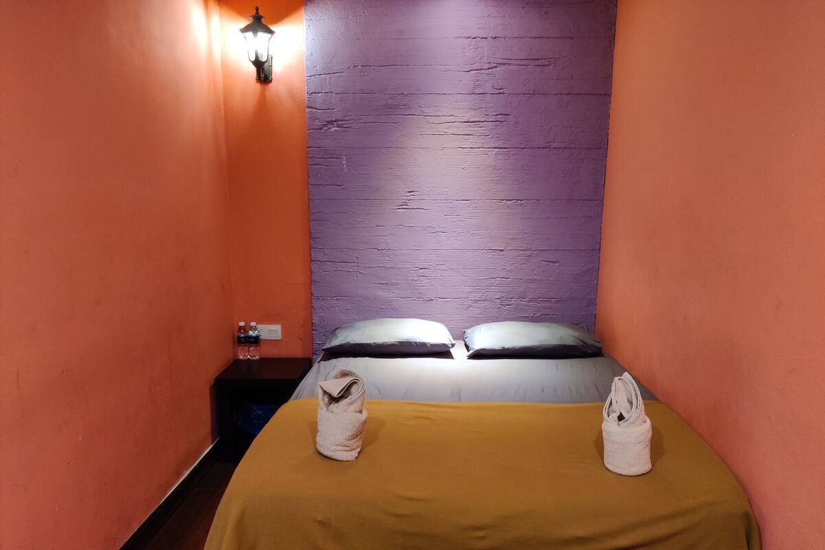 A small, narrow hotel room with a bed, two pillows, a nightstand with a lamp, and two towels on the bed.