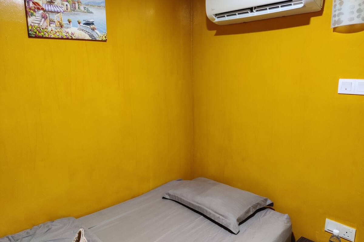 A small, yellow-walled room with a single bed, an air conditioner, and a wall-mounted power outlet.