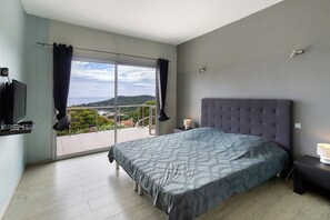A bedroom with a large bed, a television on a shelf, a nightstand with a lamp, and a sliding glass door leading to a balcony.