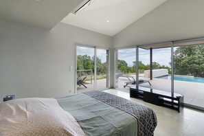 A bedroom with a large bed, a sliding glass door leading to an outdoor area with a pool and lounge chairs.