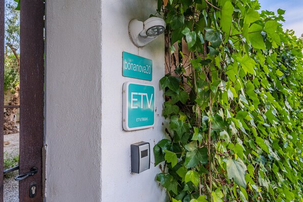 Una pared con una cámara de seguridad, una puerta con cerradura y dos letreros que indican 'bonanova20' y 'ETV' con un número de contacto.
