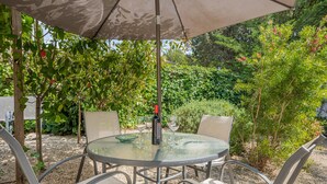 Una mesa redonda de vidrio con sillas de metal, una sombrilla, y una botella de vino, ubicada en un jardín con follaje y suelo de grava.
