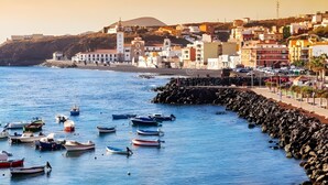 Un pueblo costero con barcos amarrados a la orilla, un rompeolas rocoso y edificios con una mezcla de estilos arquitectónicos.