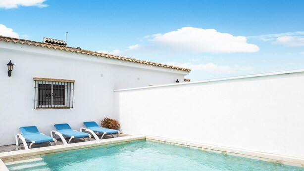 Una piscina con sillas reclinables azules, un edificio blanco con techo de tejas y un cielo azul despejado.