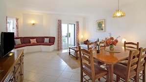 A living room with a sofa, dining table, chairs, a TV, and a lamp. - Balaia Apartment near the Beach with Shared Pools (Albufeira)