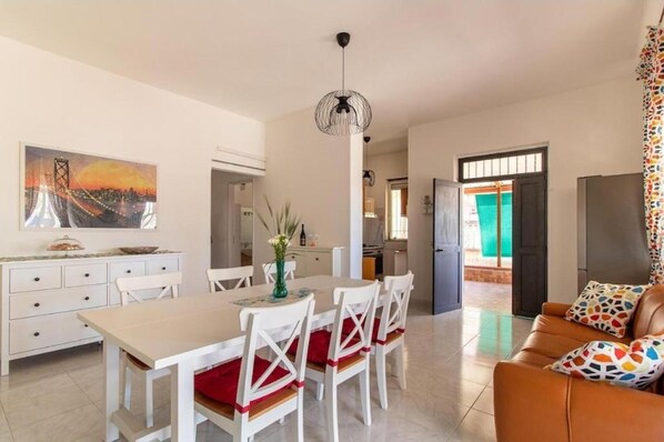 A dining area with a table and chairs, a sofa, a painting, and a kitchen in the background.