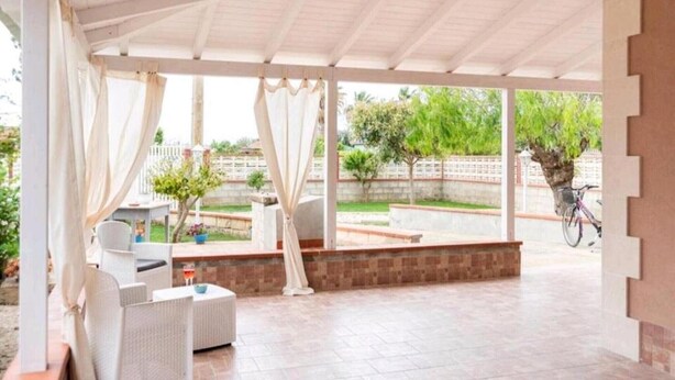 Un área de estar techada al aire libre con mobiliario de mimbre y vistas a un jardín.
