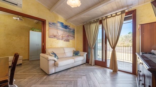 Una sala de estar con un sofá, piso de madera, un ventilador de techo y vistas a través de una ventana con cortinas.