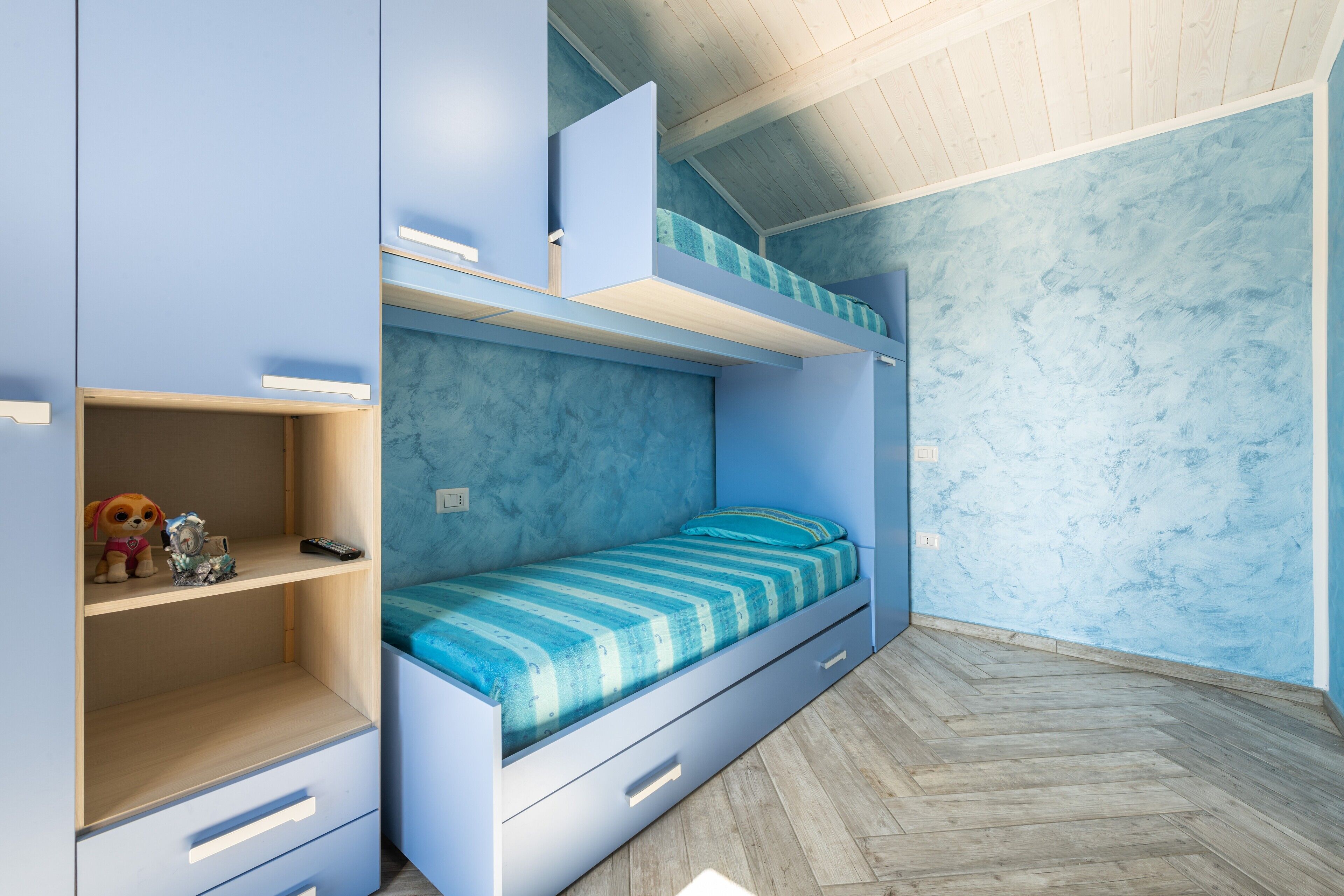 Ein Etagenbett mit blauer Kopfteil und Schubladen, ein kleines Regal mit Spielzeug und ein Parkettboden mit Fischgrätmuster.