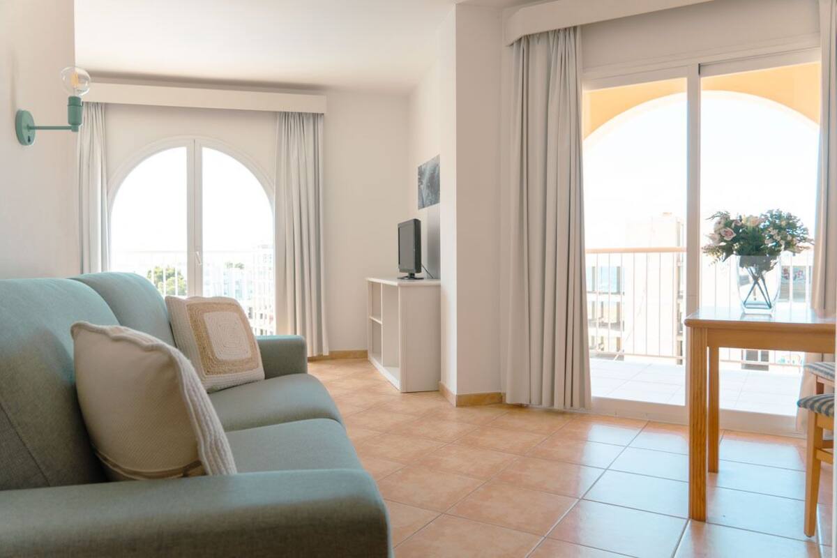 Un salon comprenant un canapé, un téléviseur posé sur un meuble, une table à manger avec des chaises et de grandes fenêtres avec des rideaux.