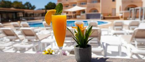 A glass of orange juice with a mint leaf garnish on a table by a poolside with lounge chairs and a potted plant.