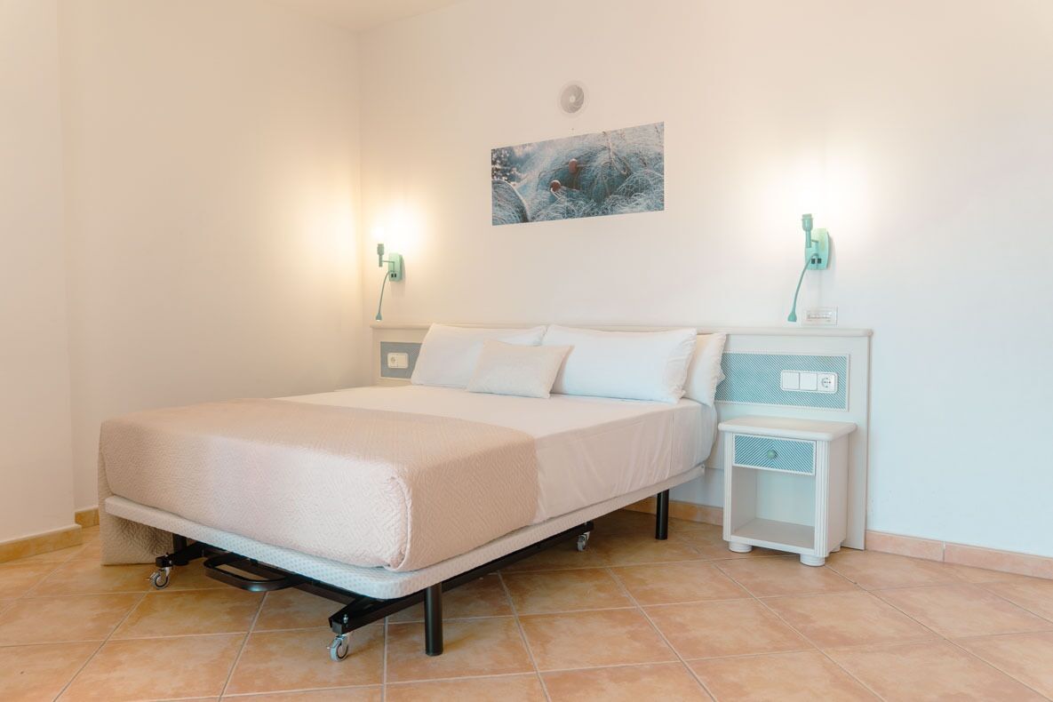 A single-bed room with a mattress on wheels, a bedside table with a lamp, and a wall-mounted artwork.
