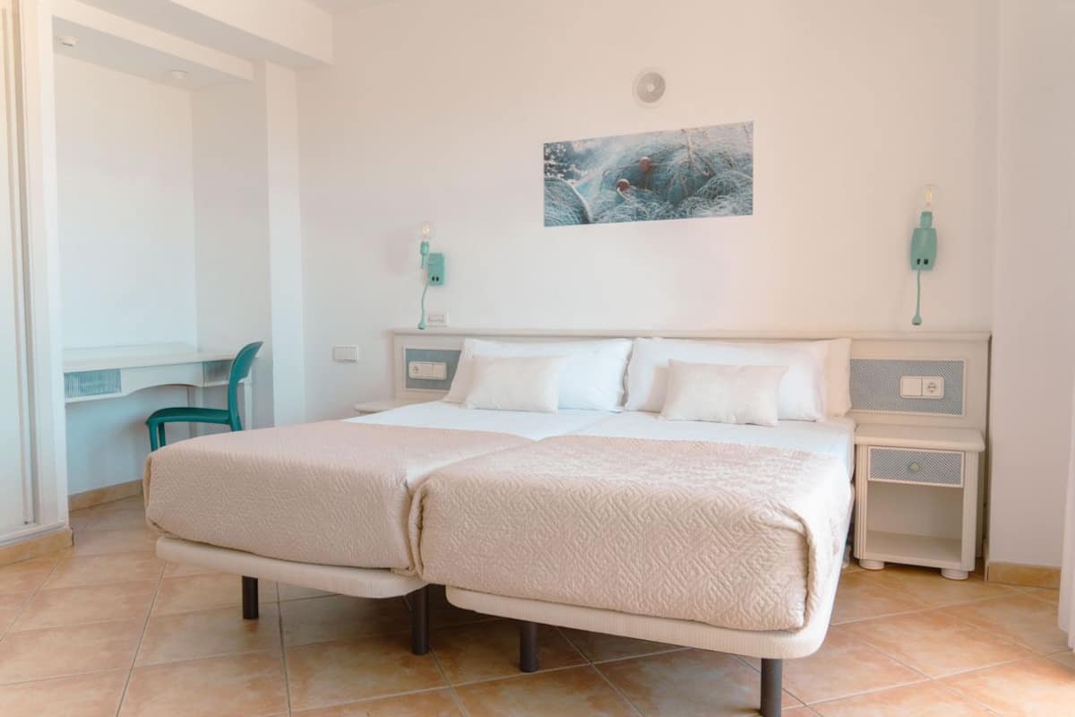 Un lit double avec une couette beige, un bureau avec une chaise de couleur turquoise et une œuvre d’art accrochée au mur.