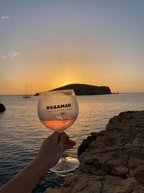 A hand holding a glass of rosé wine with the label 'Rosamar Ibiza' against a sunset backdrop with a body of water and a distant landform. - Rosamar Ibiza Hotel (Sant Josep de sa Talaia)