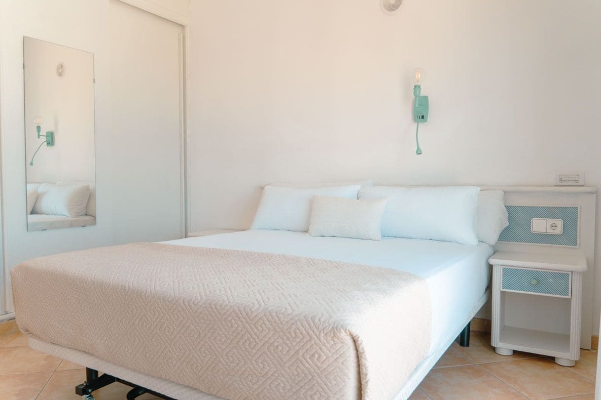 A neatly arranged bedroom with a large bed, a bedside table, a nightstand, and a wall-mounted light fixture.