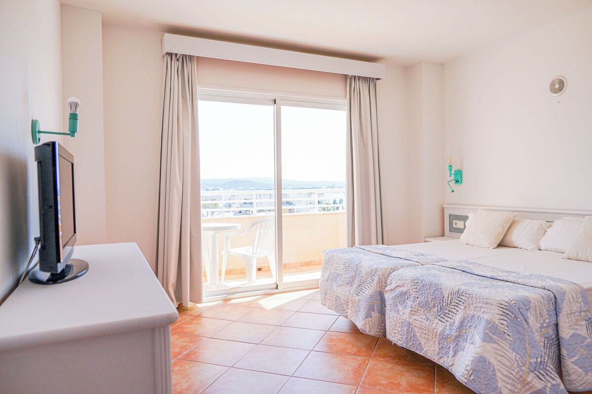 A hotel room with a bed, a TV on a stand, a balcony with a view of the city, and a window with curtains.