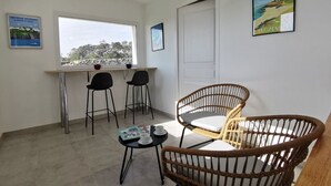 A room with a large window, a dining table with chairs, a wicker chair, and a small table with a magazine and cups.