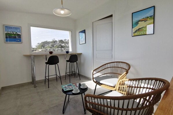 Una habitación con un ventanal grande, una mesa de comedor con sillas, una silla de mimbre y una mesita con una revista y tazas.