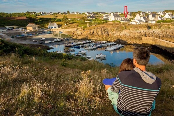 Un paisaje costero con un puerto deportivo lleno de barcos, una pareja sentada en una colina cubierta de hierba con vistas al agua, y casas al fondo.