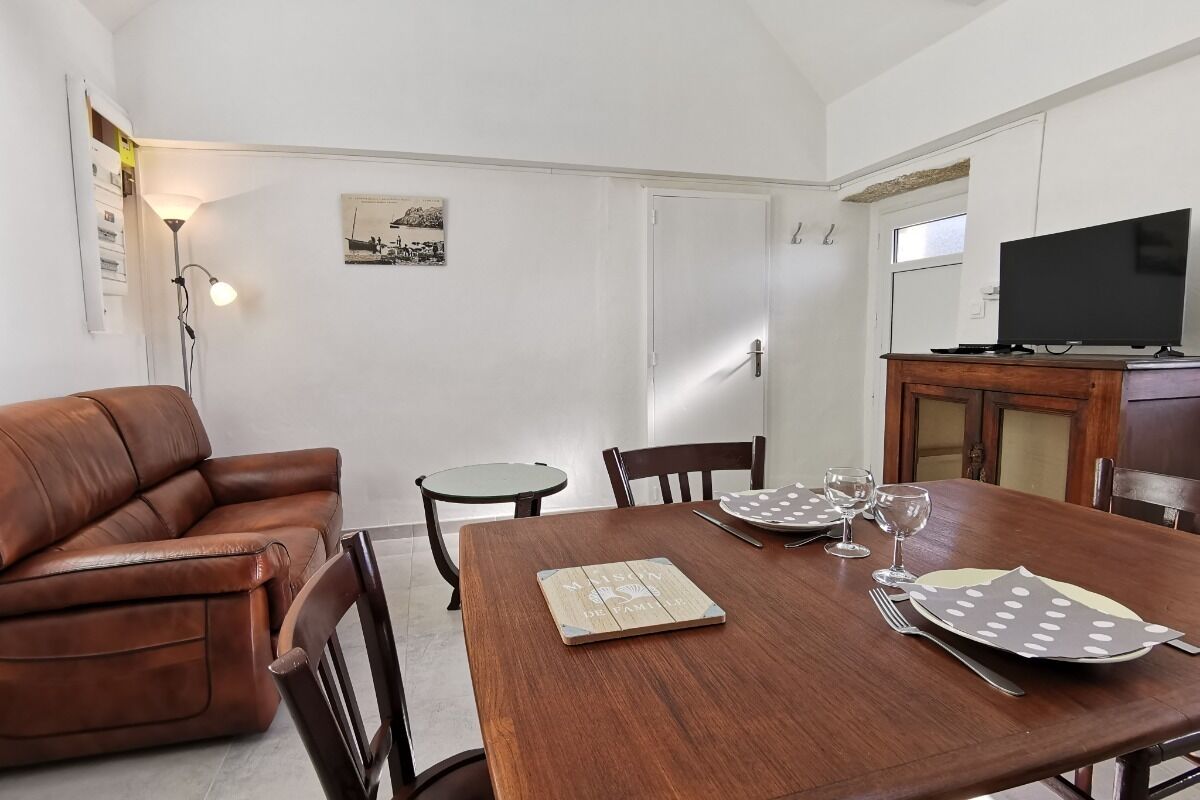 A dining area with a wooden table set for two, a brown leather couch, and a television on a wooden cabinet.