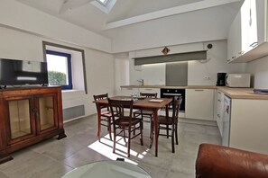 A modern kitchen with a dining table and chairs, a TV on a wooden cabinet, and a window with blinds. - Maison de Pêcheur Avec vue Imprenable sur la mer et le Port (Plozévet)