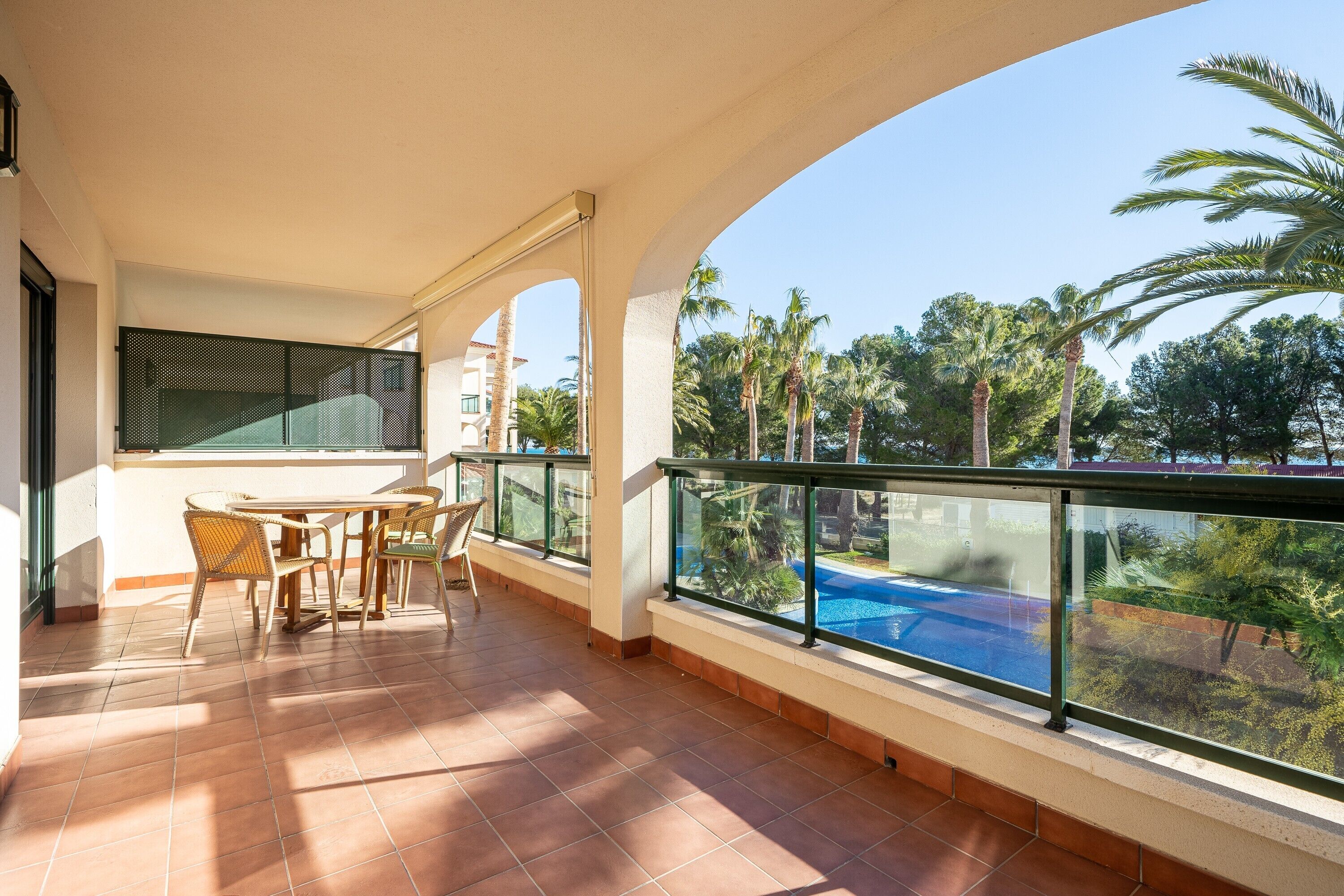 A balcony with a table and chairs, a pool, and palm trees in the background.