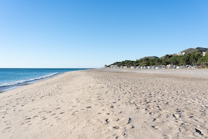 A wide sandy beach with footprints, a clear blue sky, and buildings in the background.