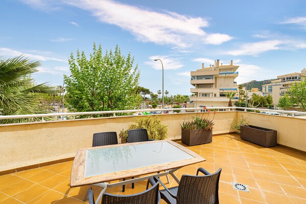 A rooftop terrace with a glass table, wicker chairs, potted plants, and a view of buildings in the background.