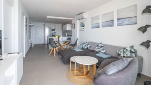 A modern living room with a grey sofa, a small white table, and a kitchen in the background. - 'Apartment ARCE Frontline Seaview' with Pool, Balcony & Wi-Fi (Nerja)