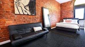 A room with a brick wall, a black leather sofa, a bed with white bedding, and a large mirror. - Bayside House - Hostel (St Kilda)