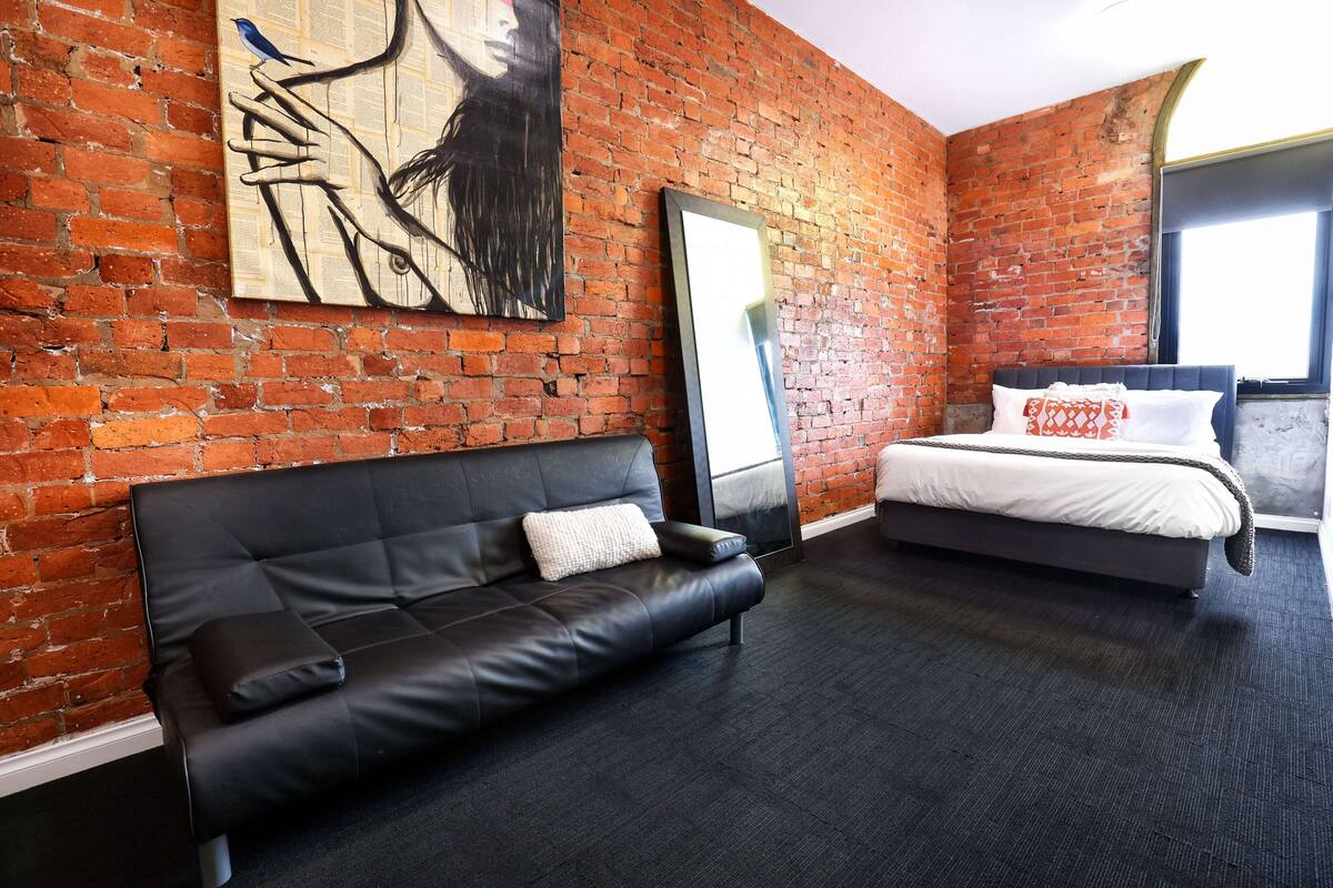A room with a brick wall, a black leather sofa, a bed with white bedding, and a large mirror.