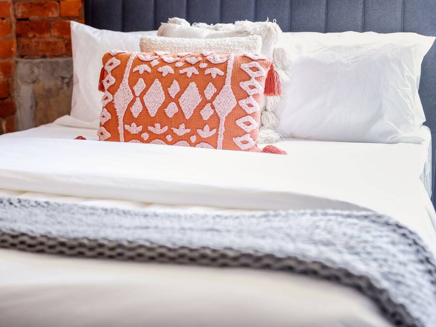 A neatly made bed with white bedding and a patterned orange throw pillow.