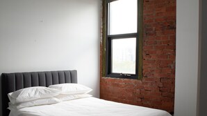 A bed with white bedding and a grey headboard in a room with a brick wall and a window. - Bayside House - Hostel (St Kilda)