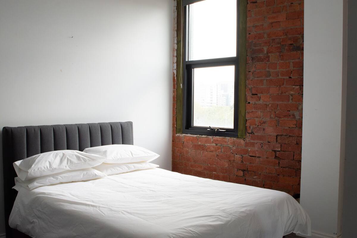 A bed with white bedding and a grey headboard in a room with a brick wall and a window.