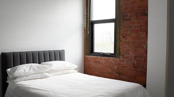 Una cama con ropa de cama blanca y un cabecero gris en una habitación con pared de ladrillo y ventana.