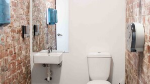 A bathroom with a brick wall, a white sink, a toilet, and a window. - Bayside House - Hostel (St Kilda)