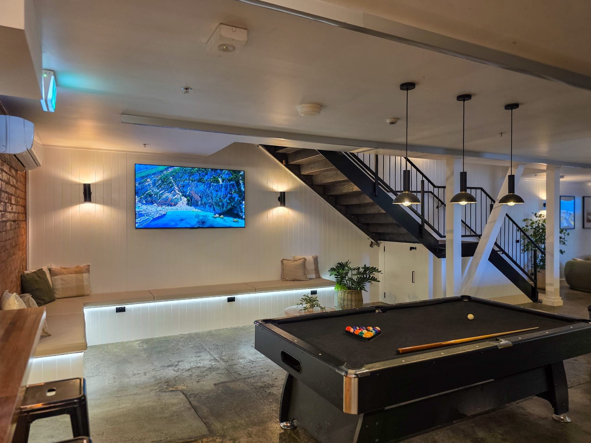 A modern living room with a pool table, a large flat-screen TV, and a staircase.
