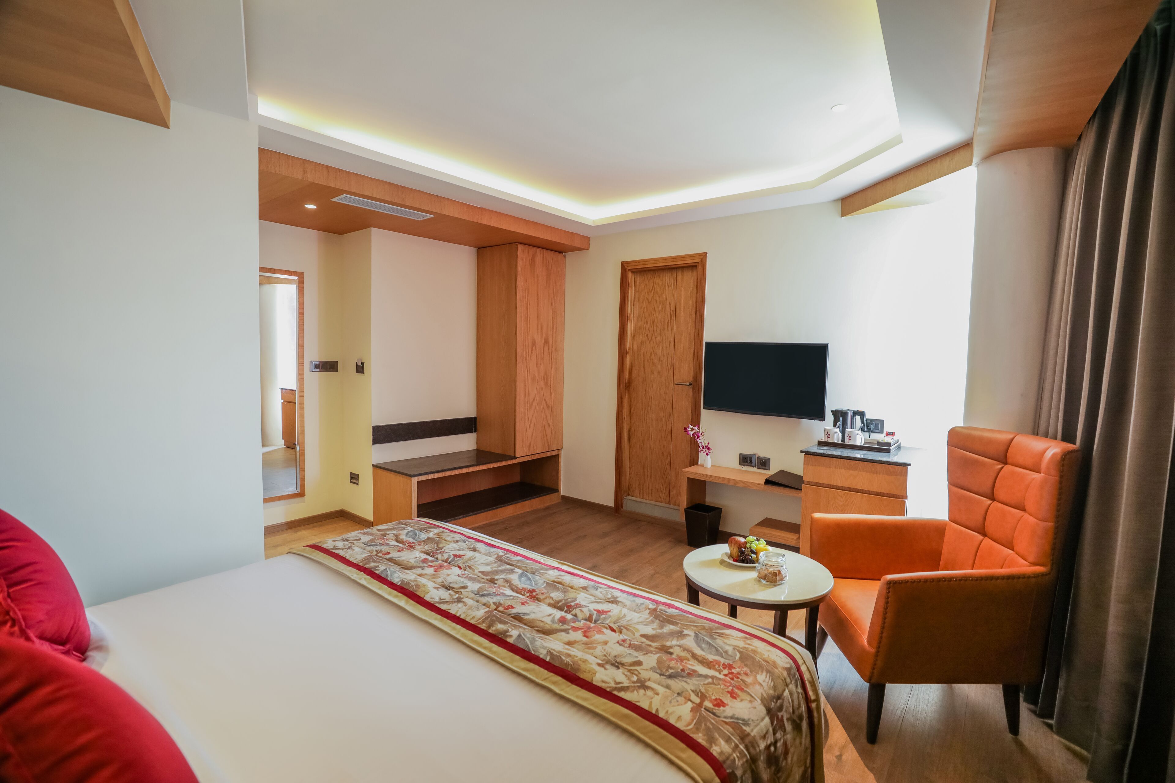 A hotel room with a bed, a small table, an orange armchair, a TV, and a wooden wardrobe.