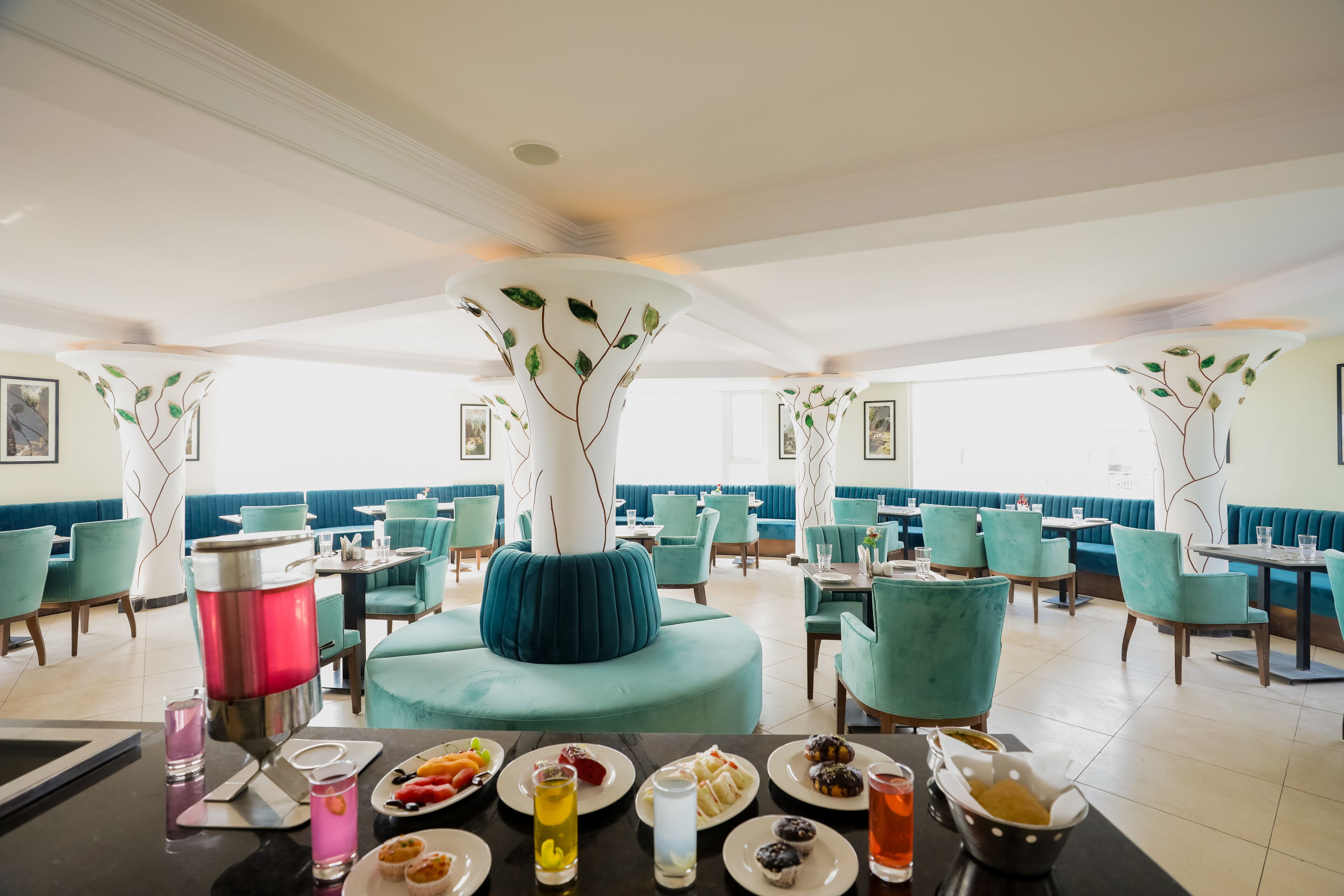 Un restaurante con sillas color turquesa, un área central de asientos acolchados azules y una variedad de platos y bebidas sobre las mesas.