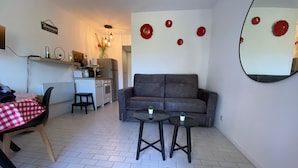 Un espacio habitable compacto con kitchenette, un sofá gris, dos mesitas pequeñas y una zona de comedor con mantel a cuadros rojo y blanco.